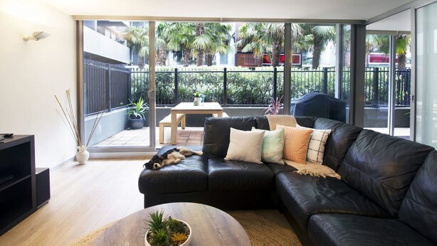 A modern living room with a black leather sofa, a wooden coffee table, and a large sliding glass door leading to a balcony with plants and a view of the neighborhood. - 250m from the beach.Gym/lap pool access,Parking, pet friendly, outdoor area, BBQ (Port Melbourne)