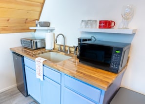 A compact kitchen with a microwave, toaster, sink, and a small shelf with mugs and a glass.