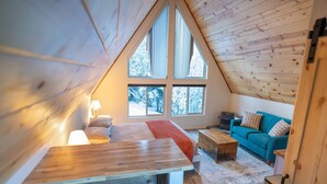 A cozy attic bedroom with a wooden ceiling, a large window, a bed, a table, and a blue sofa.