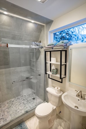 A modern bathroom with a glass shower enclosure, a toilet, a sink, and a built-in shelf with towels and toiletries.