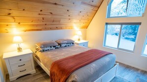A bedroom with a wooden ceiling, a bed with a red blanket, two bedside lamps, and a window with a snowy view.
