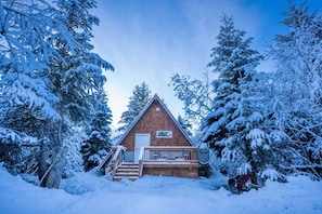A cabin with a wooden deck, surrounded by snow-covered trees.
