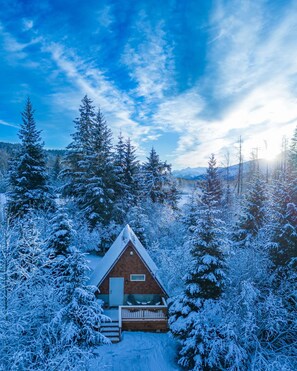 Une cabane entourée d’arbres couverts de neige, sous un ciel dégagé, avec des montagnes qui se dessinent à l’horizon.