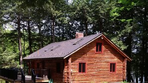 Un chalet rustique en rondins de bois, doté d’un porche, et entouré d’un paysage naturel composé de rochers et de verdure.