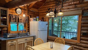L’intérieur d’un chalet en bois comprend une cuisine, une table à manger, des chaises, un réfrigérateur, une horloge et une fenêtre donnant sur la forêt.