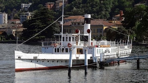 Un barco blanco amarrado en un muelle con una franja roja a lo largo del casco, rodeado por una ladera boscosa con edificios.