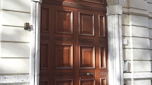 Una gran puerta de madera ornamentada con un arco decorativo encima, en un edificio con paredes de piedra clara y una barandilla de piedra.