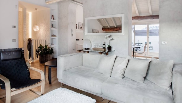 Una sala de estar moderna con un sofá blanco, una silla negra y una alfombra blanca. Hay una estantería, una mesa con una lámpara y una ventana con vistas al exterior.