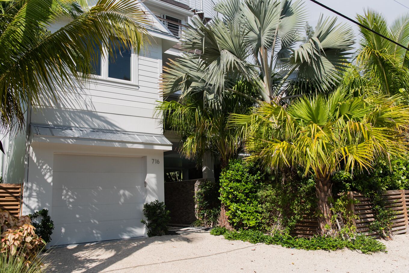 A two-story house with a white garage door, surrounded by lush tropical plants and palm trees.