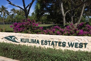 Een bordje voor Kuilima Estates West, met een logo, omringd door weelderige begroeiing en paarse bloemen.