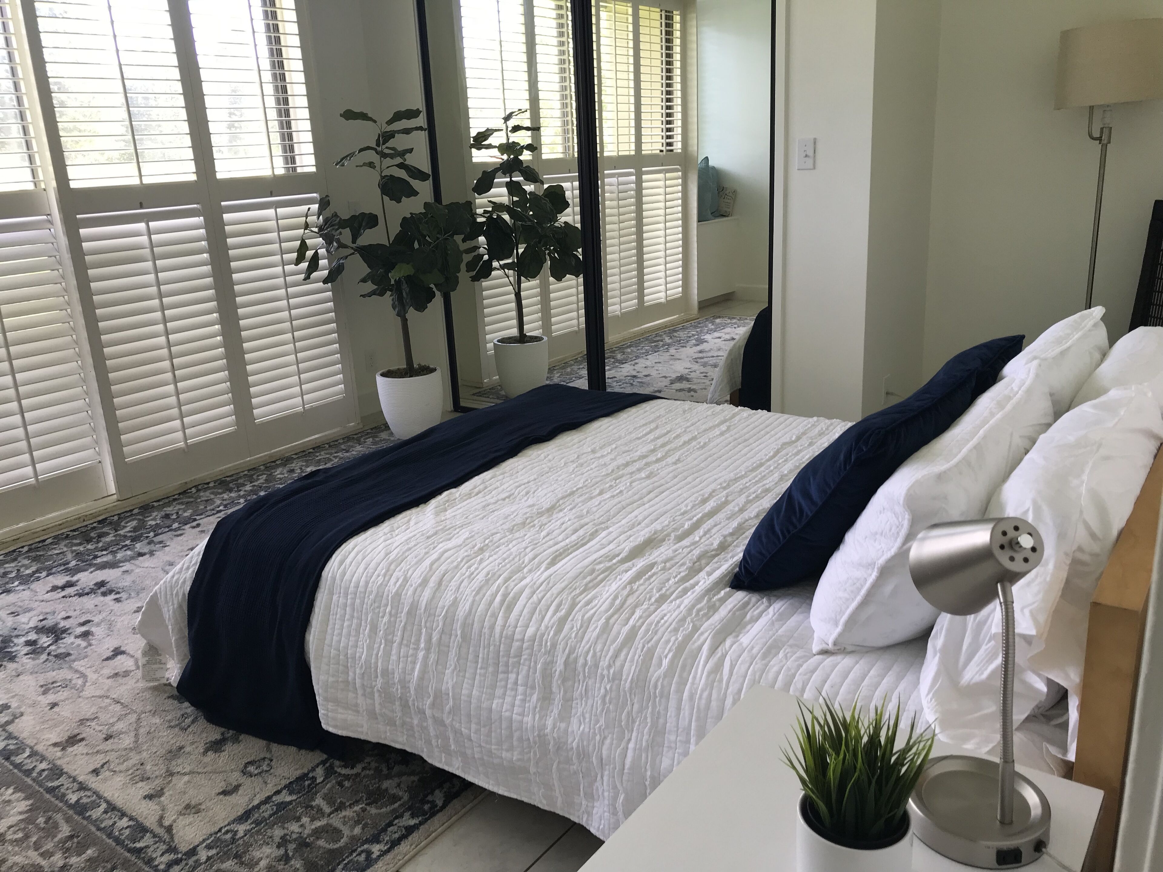 Une chambre à coucher avec un lit, une lampe de chevet, une plante en pot et une fenêtre avec des stores.