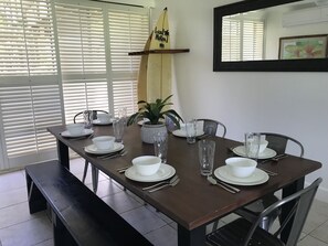 Een eettafel gedekt met witte borden, kommen en bestek, met aan weerszijden stoelen, en een potplant en een surfboard op een plank in de achtergrond.
