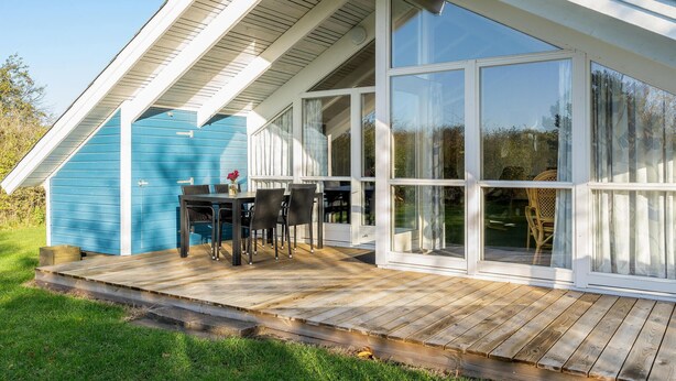 A covered patio with a wooden deck, a table with chairs, and a blue building in the background. - Gorgeous home in Hemmet with sauna (Hemmet)