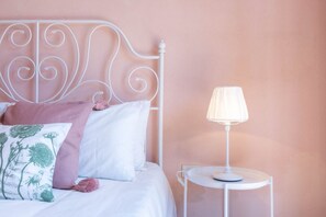 A neatly made bed with white and pink bedding, a white metal headboard, and a bedside table with a lamp.