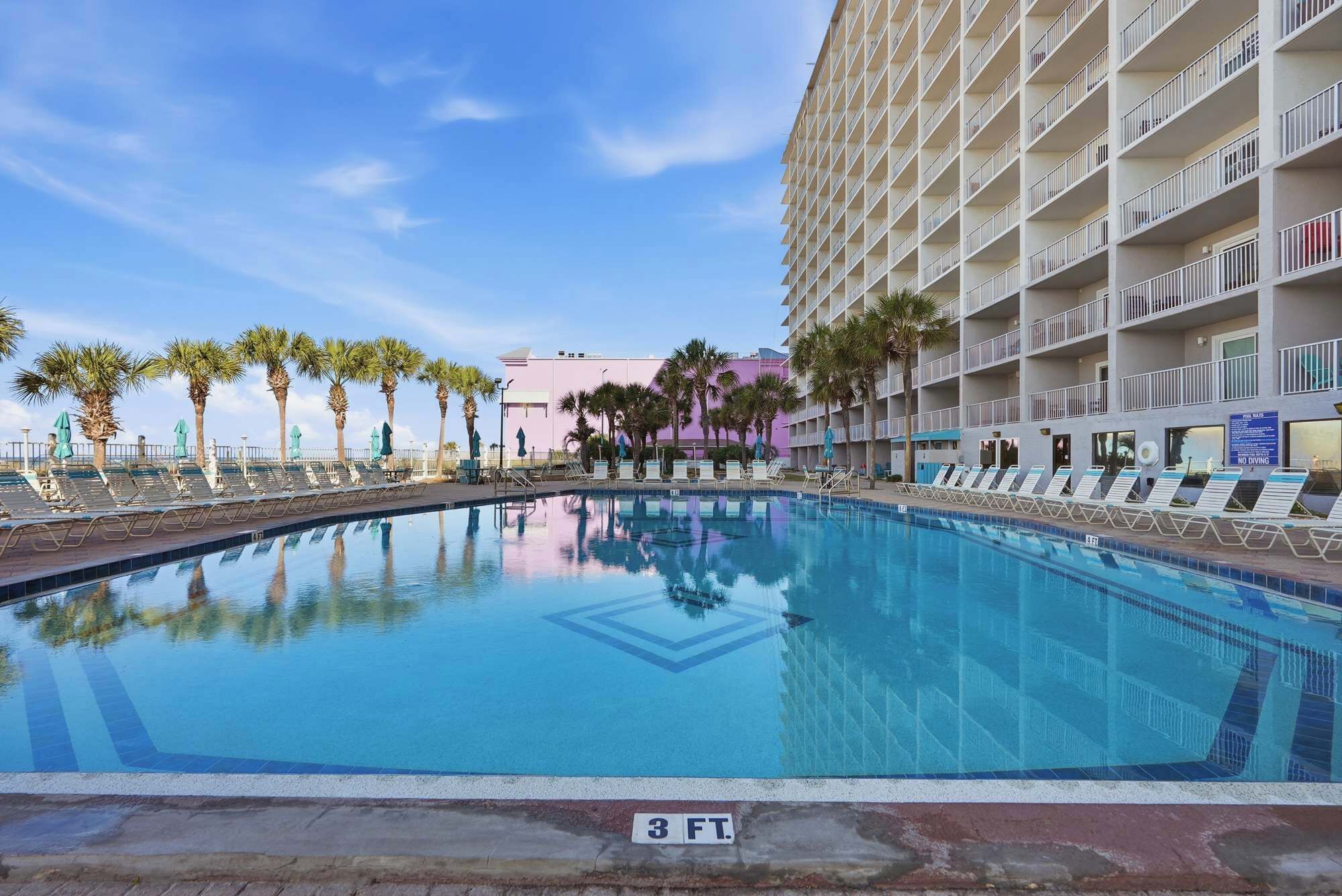 A pool with lounge chairs, palm trees, and a hotel building in the background.