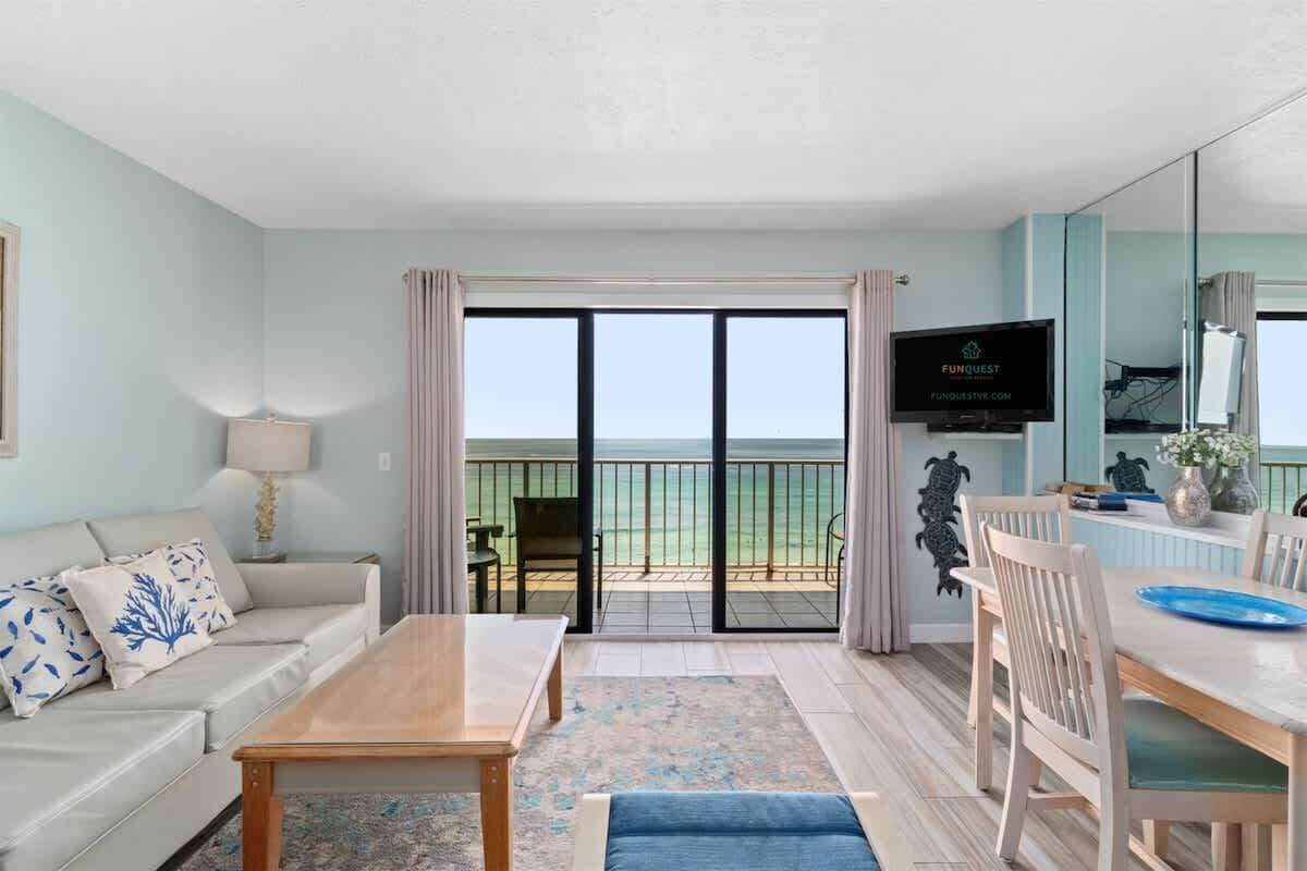 A living room with a sofa, coffee table, and dining table. There is a TV on the wall and a view of the ocean through large windows.