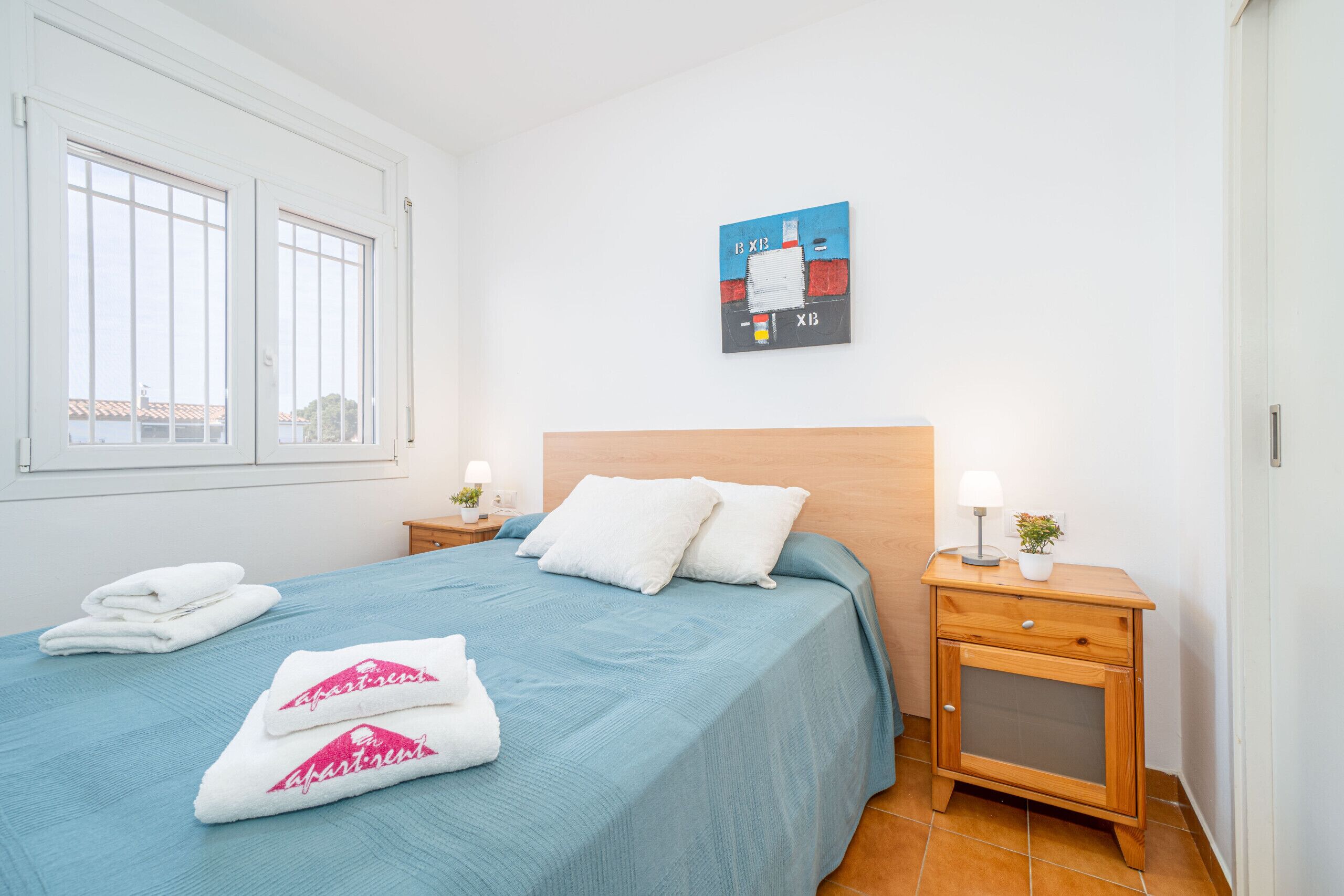 A neatly made bed with blue bedding, white pillows, and two towels with 'apartment' text. There is a small bedside table with a lamp and a plant. A painting hangs on the wall above the bed.