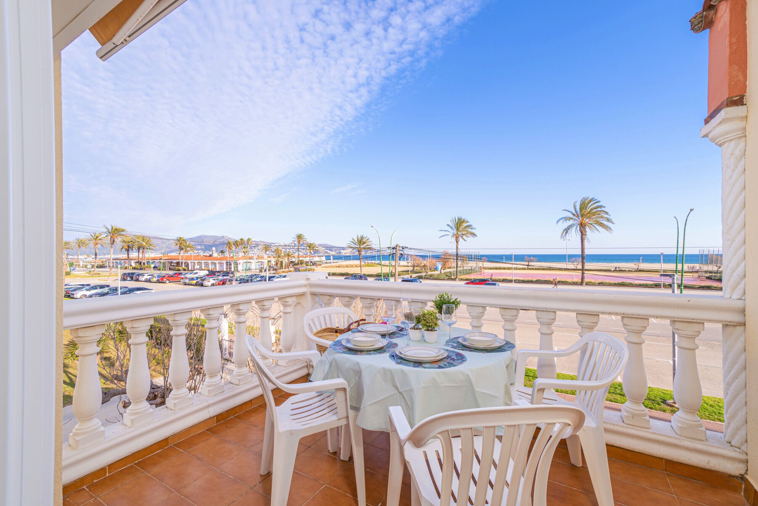 A balcony with a table set for dining, overlooking a beach with palm trees and a parking area.