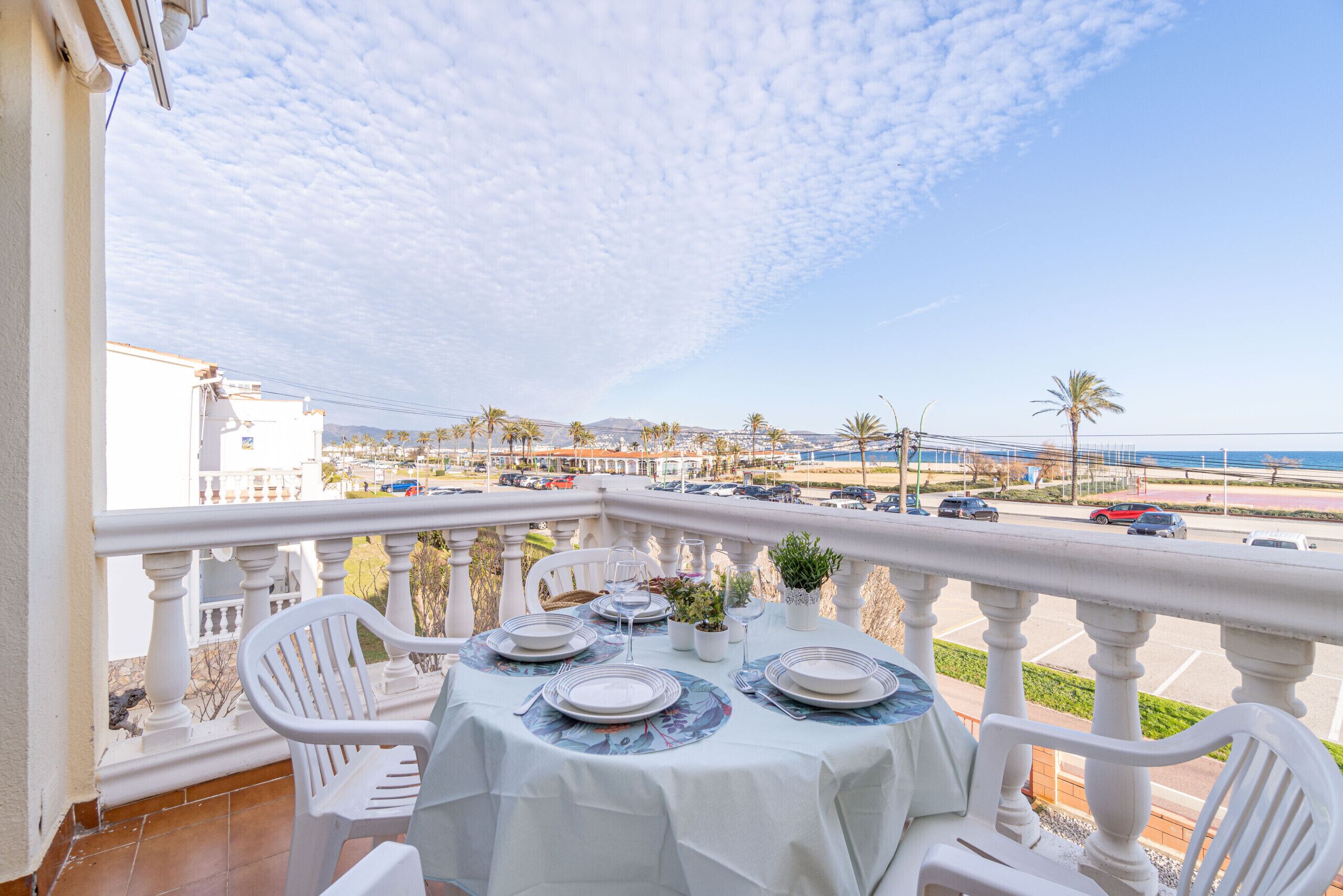 A balcony with a table set for dining, overlooking a parking area and a coastal view.