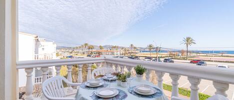 A balcony with a table set for dining, overlooking a parking area and a coastal view.
