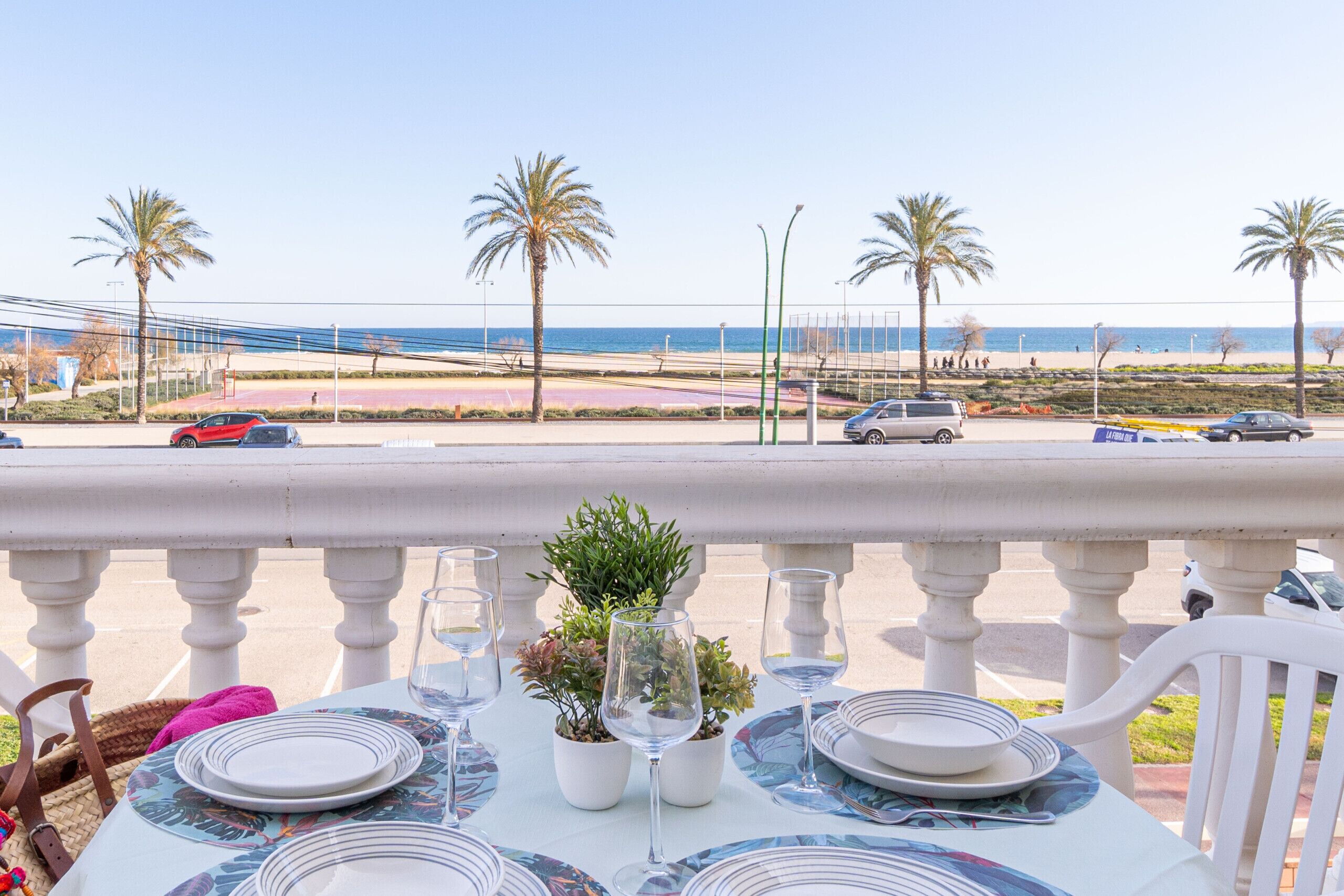 A table set with plates, glasses, and a potted plant on a balcony overlooking a beach with palm trees and a road with cars.