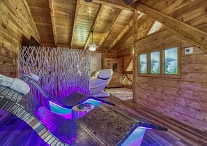 A wooden cabin with a massage chair, a small pool area with LED lighting, and a view of the mountains through the windows.