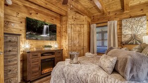 Una camera da letto accogliente in legno con camino, un letto con coperta soffice, una TV a parete che mostra una cascata e un grande quadro raffigurante un albero.