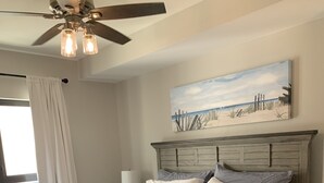 A bedroom with a wooden bed, a ceiling fan, and a painting of a beach scene. - The Club at Mexico Beach - Our Happy Place (Mexico Beach)