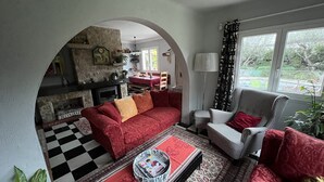 En hyggelig stue med en rød sofa, en lænestol med et mønster, og et ternet gulv. En pejs ses i baggrunden, og der er et spiseområde med et bord og stole.
