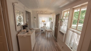 A narrow hallway with a wooden floor, a white dresser, a chair, a small table with a lamp, and a window with a view of greenery.