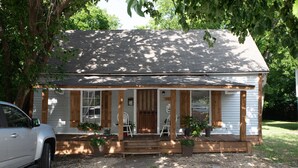 A small, single-story house with a porch, two chairs, and potted plants.