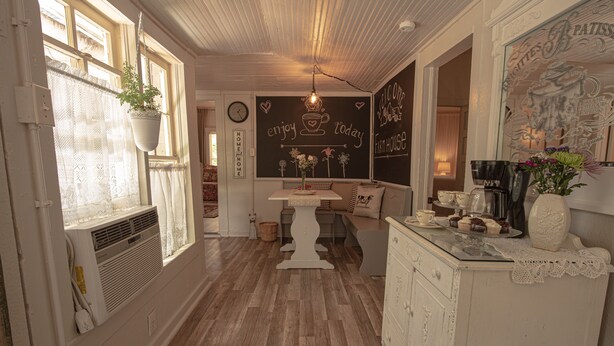 A cozy kitchen with a wooden floor, a hanging plant, a chalkboard with writing, and a dining area with a table and chairs.