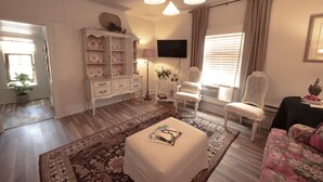 A living room with a white sofa, a white armchair, a coffee table with books, a white cabinet with dishes, a television, and a window with curtains.