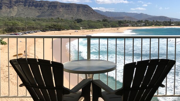 Un balcón con dos sillas y una mesa con vistas a una playa de aguas azules y montañas lejanas.