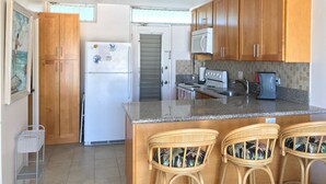 Una cocina con armarios de madera, un refrigerador blanco, un microondas, una estufa y una cafetera. Hay tres taburetes de mimbre.