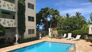 Una piscina con sillas de descanso y una estatua, rodeada de exuberante vegetación y un edificio con balcones.