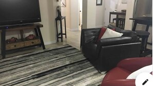Una sala de estar con un televisor de pantalla plana sobre un mueble, un sofá de cuero negro con un cojín rojo, una alfombra a rayas y una mesa de centro de madera.
