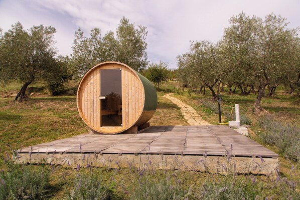 A wooden barrel structure with an open door, situated on a wooden deck surrounded by lavender fields and olive trees.