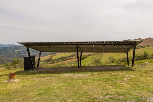 Un espacio al aire libre cubierto con techo metálico, rodeado de terreno cubierto de hierba y colinas al fondo.