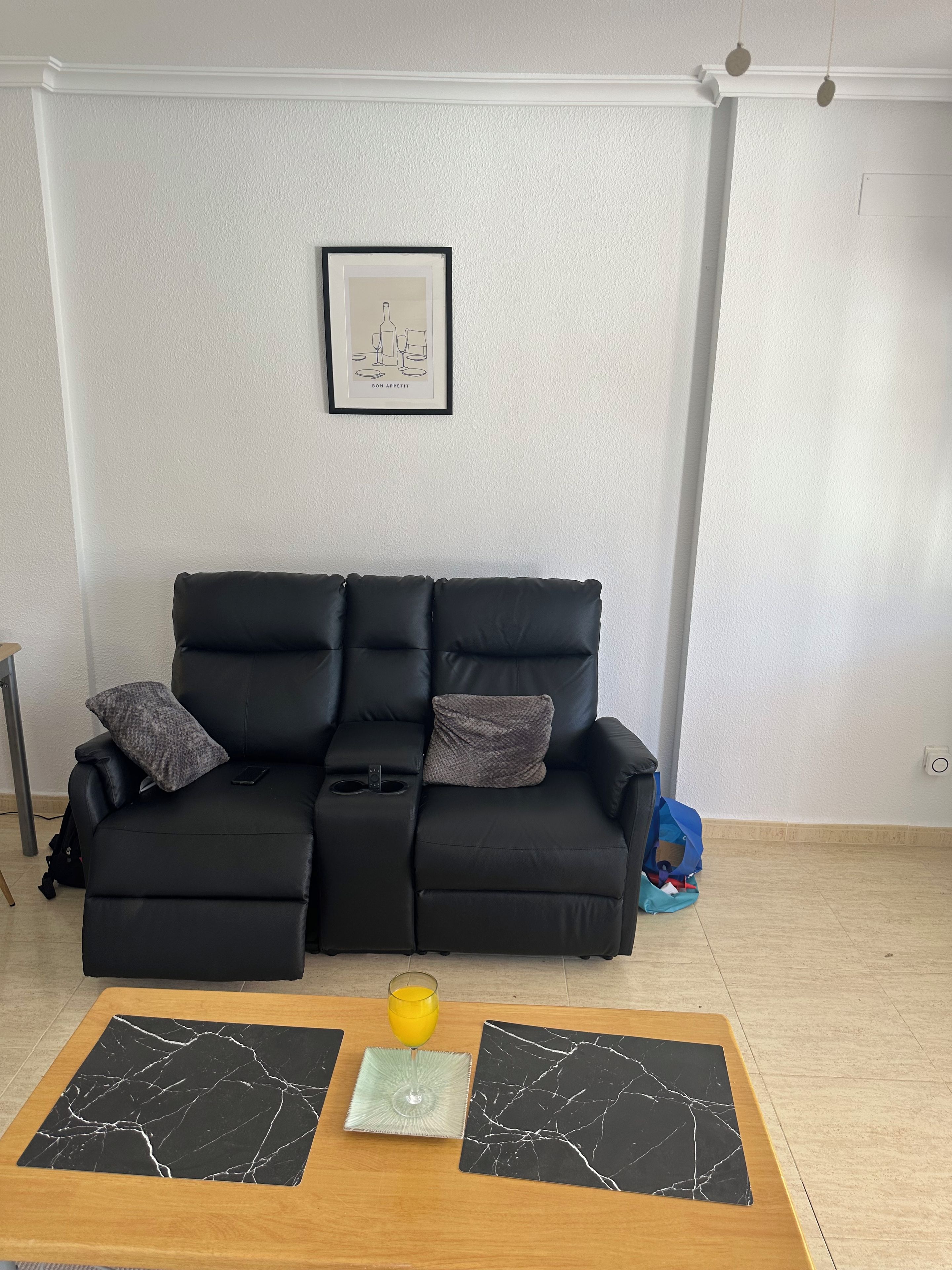 Un canapé en cuir noir avec deux coussins, un tableau encadré accroché au mur et une table basse en bois sur laquelle reposent deux sets de table et un verre de jus d’orange.