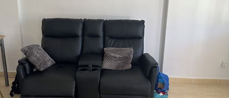 Un canapé en cuir noir avec deux coussins, un tableau encadré accroché au mur et une table basse en bois sur laquelle reposent deux sets de table et un verre de jus d’orange.