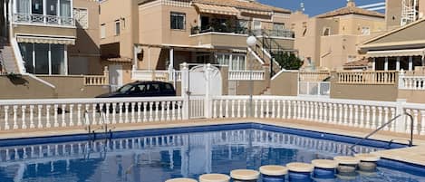 Una piscina con barandilla blanca, rodeada de edificios color beige con balcones y un cielo azul despejado.