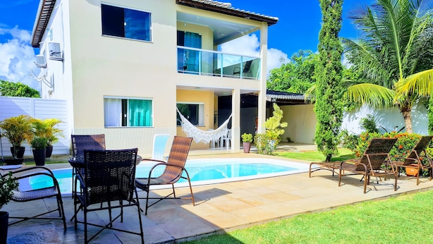 Una casa de dos pisos con piscina, mobiliario exterior y una hamaca.