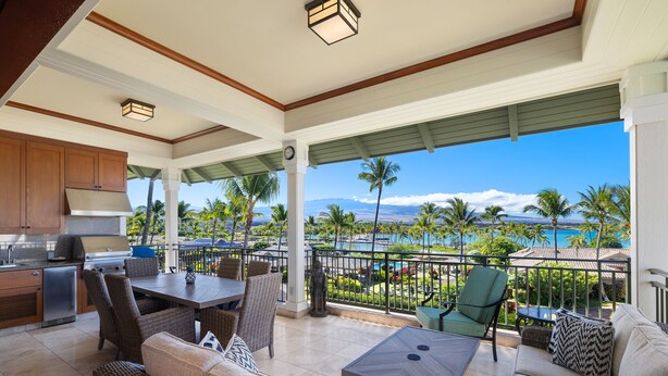 A covered outdoor area with a dining table, chairs, and a sofa. There is a kitchen area with a grill and cabinets. The view includes palm trees and a body of water. - 3 BR penthouse with easy beach access - Hilton Waikoloa Access Included (Waikoloa)