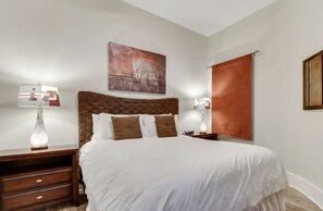 A bedroom with a large bed, two bedside tables with lamps, a wall-mounted painting, and a window with a blind. - Luxury 12th Floor Condo at Bella Luna (Orange Beach)