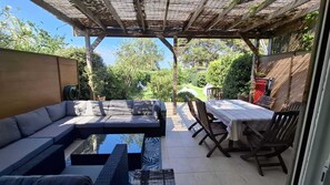 En overdækket terrasse med sofa, spisebord og stole, omgivet af grøn vegetation.