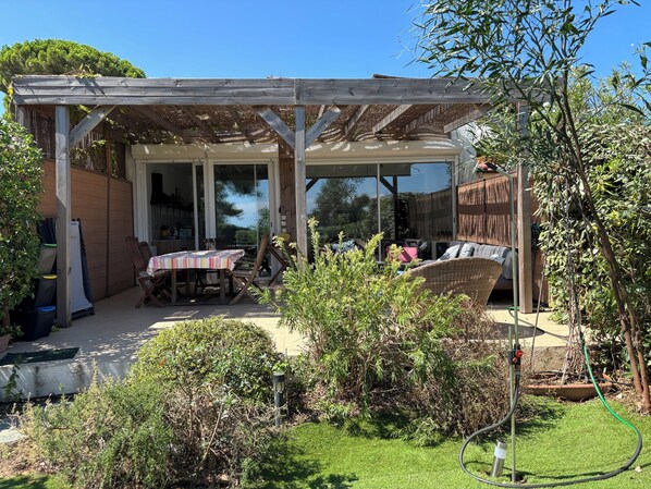 En overdækket terrasse med et bord og stole, en fletsofa og et haveområde med en haveslange.