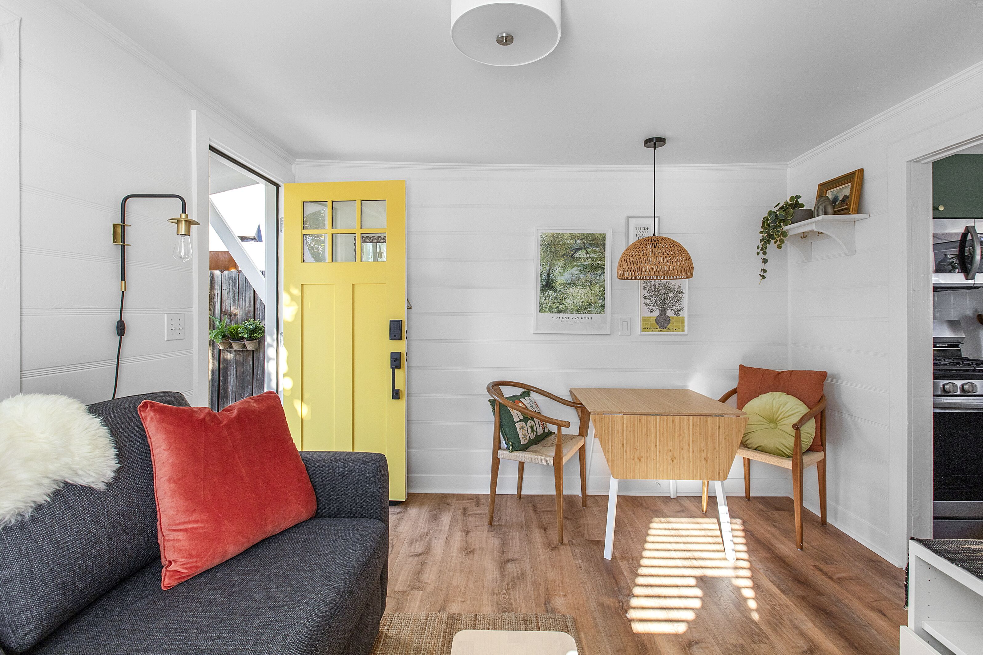 Ein modernes Wohnzimmer mit einer gelben Tür, einem grauen Sofa mit einem roten Kissen, einem Holzes Esstisch mit zwei Stühlen und einer hängenden Lampenleuchte.
