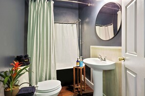 Ein Badezimmer mit Toilette, Waschbecken und Duschvorhang. Eine Topfpflanze und ein kleiner Hocker mit Pflegeprodukten sind vorhanden.