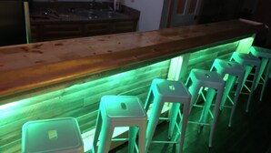 Un espace bar doté d’un comptoir en bois et d’un éclairage vert, avec plusieurs tabourets blancs.